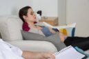 Seorang wanita berbaring di sofa dengan mata terpejam, terlihat santai, sementara orang lain duduk di dekatnya sambil memegang buku catatan dan pena, menunjukkan sesi hipnoterapi atau konseling.