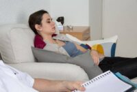 Seorang wanita berbaring di sofa dengan mata terpejam, terlihat santai, sementara orang lain duduk di dekatnya sambil memegang buku catatan dan pena, menunjukkan sesi hipnoterapi atau konseling.