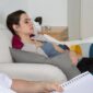 Seorang wanita berbaring di sofa dengan mata terpejam, terlihat santai, sementara orang lain duduk di dekatnya sambil memegang buku catatan dan pena, menunjukkan sesi hipnoterapi atau konseling.